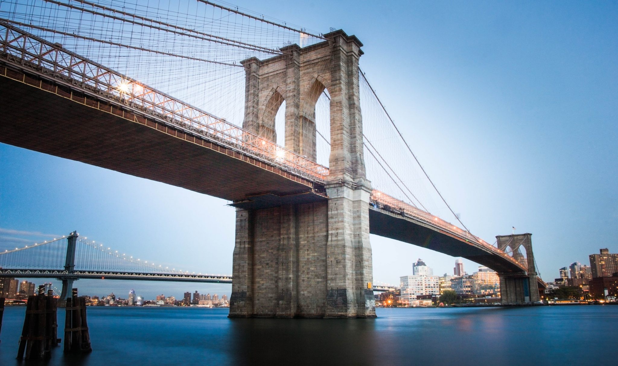 The Brooklyn Bridge at dusk, viewed from the water with its stone tower, suspension cables, and the New York City skyline in the background.