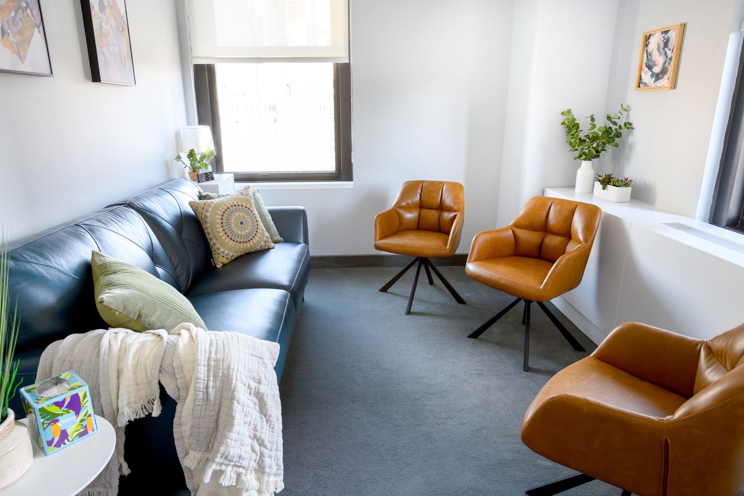 Dr. Davis's Brooklyn therapy office, with a navy leather sofa, tan leather swivel chairs, plants, and natural light from a large window.
